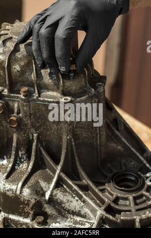 Die Hand der Mechaniker in einem schwarzen Nitril Schutzhandschuh schraubt die Schraube aus dem Gehäuse des Getriebes. Stockfoto