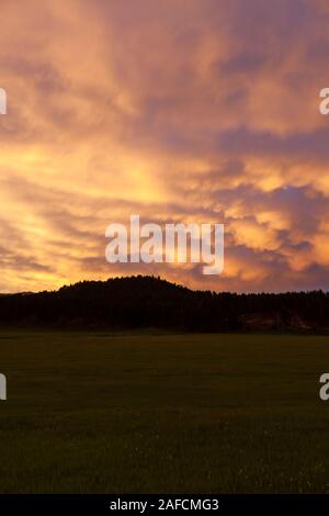 Eine dramatische gelb und rosa Sonnenuntergang auf gewitterwolken am späten Abend mit weit entfernten Bergen und einem offenen Feld reflektiert. Stockfoto