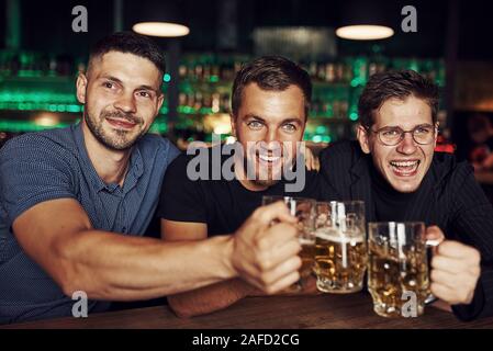 Klopfen Gläser. Drei sport Fans in einer Bar beobachten Fußball. Mit einem Bier in der Hand Stockfoto