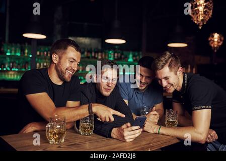 Lustige Inhalte auf dem Smartphone. Drei Freunde in der Kneipe ruht mit einem Bier in der Hand. In Gespräch Stockfoto