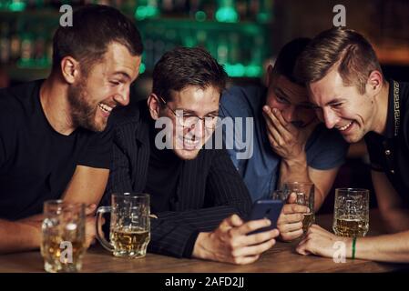 Lustige Inhalte auf dem Smartphone. Drei Freunde in der Kneipe ruht mit einem Bier in der Hand. In Gespräch Stockfoto