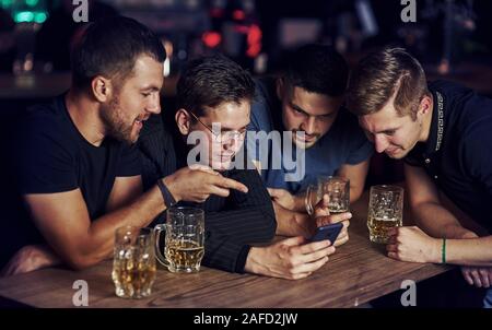 Lustige Inhalte auf dem Smartphone. Drei Freunde in der Kneipe ruht mit einem Bier in der Hand. In Gespräch Stockfoto