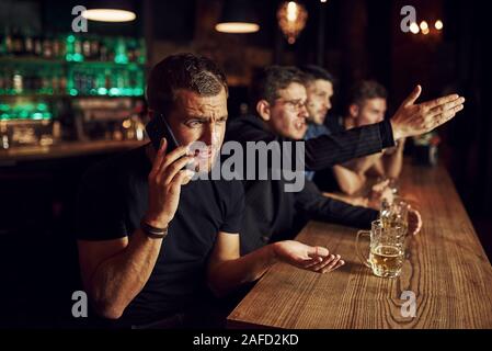 Man spricht von dem Telefon. Drei sport Fans in einer Bar beobachten Fußball. Mit einem Bier in der Hand Stockfoto