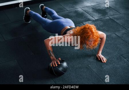 Dabei Push-ups mit Kugel. Sportliche rothaarige Mädchen haben fitness Tag in der Turnhalle am Tag. Muskulöse Körper Typ Stockfoto