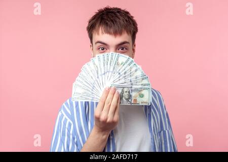 Porträt der dunkelhaarige Mann mit großen erstaunten Augen im casual Striped Shirt peeking aus Dollarscheine, seine Hälfte Gesicht verstecken, an Kamera suchen mit astoni Stockfoto