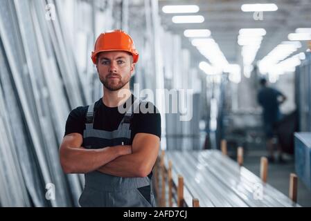 Stände mit verschränkten Armen. Portrait der männlichen Industriearbeiter im Innenbereich im Werk. Junge Techniker mit orange Schutzhelm Stockfoto