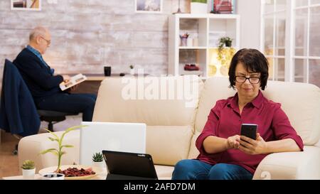 Zoom im Schoß der älteren Alter Frau mit Brille nehmen einen Schluck Kaffee surfen auf Handy. Ältere alter Mann ein Buch lesen im Hintergrund. Stockfoto