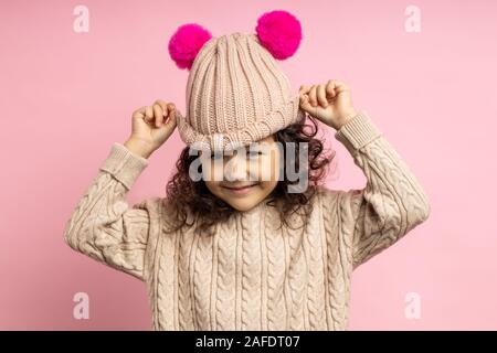 Charmantes kleines Mädchen mit lockigem Haar tragen, beige Pullover, Strickmütze mit Pink pom poms mit zufriedenen Ausdruck über rosa Hintergrund posiert. Stockfoto