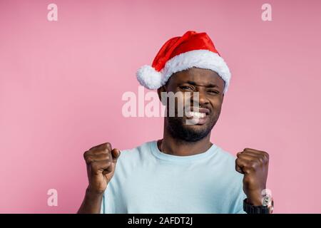 Frustriert, wütend, aggressiv afrikanischer Mann ballte die Fäuste und Zähne, Hände, tragen t-shirt, Santa hat, neben der Suche mit wütenden Ausdruck, p Stockfoto