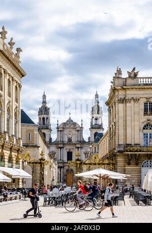 Menschen, die am Grand Hotel auf dem Stanislas-Platz in Nancy, Frankreich, vorbeischlendern und von einem vergoldeten Tor mit der Kathedrale von Nancy im Hintergrund geschlossen werden. Stockfoto