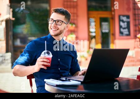 Lächelnder Mann mit Plastik Tasse frischen Saft in der Hand arbeitet mit Laptop. Geschäftsmann in Gläser Getränke trinken für Körper Flüssigkeitszufuhr während der Arbeit Stockfoto