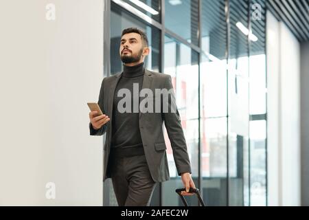 Junge Unternehmer in formale ziehen Koffer beim Verschieben in Richtung Ausgang Stockfoto