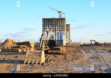 Bagger auf einer Baustelle auf dem Hintergrund einer Baukräne und Gebäude. Tieflöffel graben den Boden für die Stiftung, Festlegung Sturmabwasserkanal Stockfoto