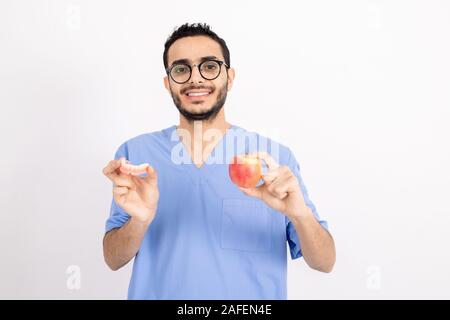 Lächelnden jungen Zahnarzt in der blauen Uniform zeigen Ihnen falsche Zähne und reifer Apfel Stockfoto