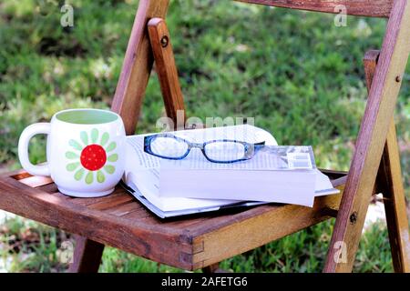 Bücher auf Holzstäbchen Stuhl in dappled Schatten mit Brille und Kaffeebecher. Stockfoto