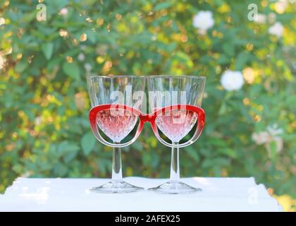 Eine skurrile Einstellung von zwei Weingläser mit Rose Wein und rote Brillen steht vor einem Rosenbusch. Stockfoto