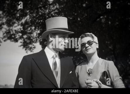 1960er Jahre, historisch, ein junges erwachsenes Paar, das draußen in gehobener Mode stand, mit dem Mann in einem Oberteil, Hut und Schwanz, mit der jungen Dame in einem Kleid und modischem Hut, Dublin, Irland. Stockfoto