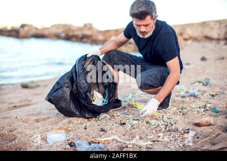 Mann in dunklem Shirt und Shorts mit weißen Handschuhen und großen schwarzen Paket Garbage Collecting am Strand. Umweltschutz und Planeten Verschmutzung co Stockfoto