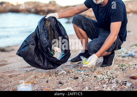 Mann in dunklem Shirt und Shorts mit weißen Handschuhen und großen schwarzen Paket Garbage Collecting am Strand. Umweltschutz und Planeten Verschmutzung co Stockfoto