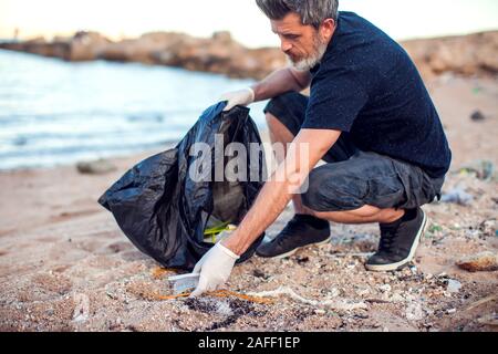 Mann in dunklem Shirt und Shorts mit weißen Handschuhen und großen schwarzen Paket Garbage Collecting am Strand. Umweltschutz und Planeten Verschmutzung co Stockfoto