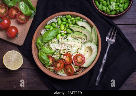 Eine frische, gesunde Salate mit zoodles Zucchini Nudeln, baby Tomaten, Avocado und Edamame Bohnen. Der Salat ist in eine hölzerne Schüssel auf eine dunkle Bambus Tabelle w Stockfoto