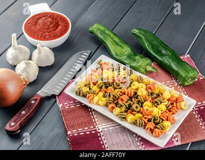 Platte von bunten rohe Nudeln, mit seinen bereit, Tomaten, Zwiebeln, Knoblauch und Paprika zum Schneiden und Kochen Stockfoto