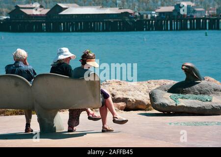 Die Leute sitzen auf der Whale Tale Bänke mit Blick auf den Hafen von Santa Barbara mit einer Bronze dolphin Skulptur und Sterns Wharf, Santa Barbara, CA, USA Stockfoto