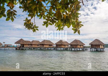 Blick auf eine Reihe von typischen strohgedeckten Beachside Überwasser Bungalows durch einen von Palmen gesäumten Strand auf Bora Bora, einer kleinen Insel im Südpazifik in Französisch-Polynesien Stockfoto