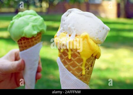 Eis hautnah. Weiß, Gelb und Grün Eis in der Waffel Kegel in der Natur. Weibliche hand mit Eis in der Waffel Kegel auf ein verschwommenes Grün Stockfoto