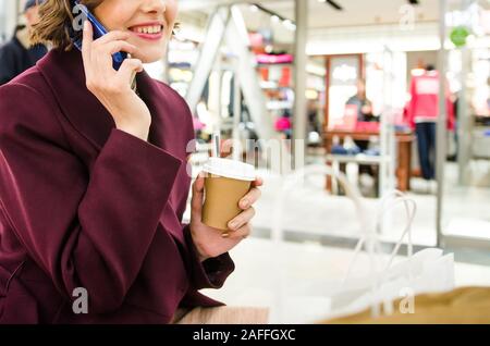 7/8 Schuß einer hübschen Frau. Shopping in der Mall. Lächelnd attraktive Frau in lila Mantel mit Coffee to go Stockfoto