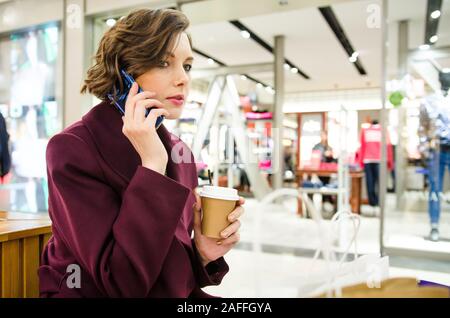 7/8 Schuß einer hübschen Frau. Shopping in der Mall. Lächelnd attraktive Frau in lila Mantel mit Kaffee trinken gehen und Telefon Stockfoto