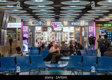 Passagiere sitzen auf Sitzen vor einem Tech & Technik Shop im Terminal 4 S von Adolfo Suárez Flughafen Barajas, Madrid, Spanien Fliegen Stockfoto