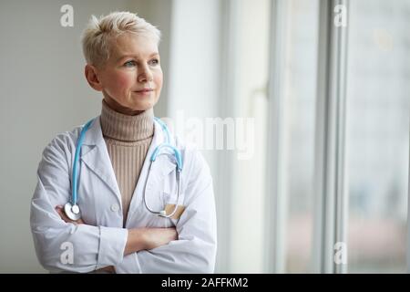 Taille bis Portrait von reife weibliche Arzt stehend mit Waffen durch Fenster in Klinik gekreuzt und weg schauen nachdenklich, kopieren Raum Stockfoto