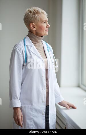 Taille bis Portrait von reife weibliche Arzt stehen durch Fenster in Klinik und weg schauen nachdenklich Stockfoto