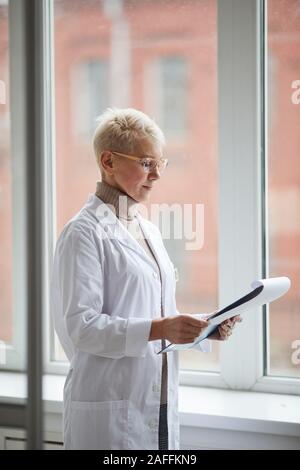 Seitenansicht Portrait von reife weibliche Arzt holding Zwischenablage beim Stehen durch Fenster in Klinik und Patient Formen, kopieren Raum Stockfoto