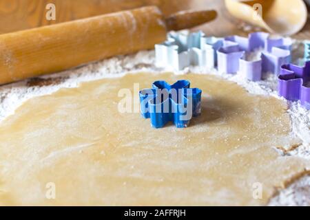 Blaue Blume Cookie Cutter Ein ausgerollt Zucker Plätzchenteig. Zusätzliche Schneeflocke und Blume Ausstecher unscharf im Hintergrund. Holz- Rollen Stockfoto