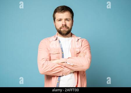 Ruhe, bärtigen Mann steht selbstbewusst und mit verschränkten Armen und starrt angestrengt in die Kamera. Auf einem blauen Hintergrund. Stockfoto