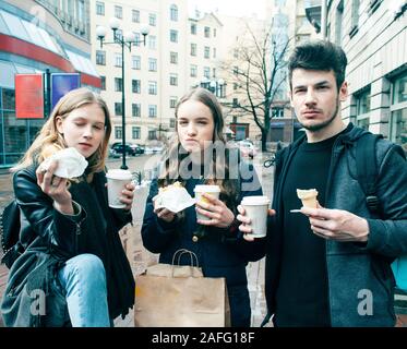 Lifestyle und Personen Konzept: zwei Mädchen und Guy essen fast food auf Stadt Straße Zusammen Spass haben, trinken Kaffee Stockfoto