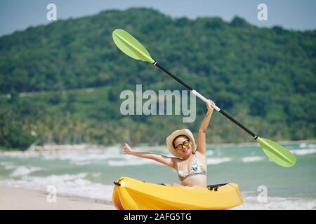 Freudige junge vietnamesische Frau in Hut und Sonnenbrille in Kajak Boot sitzend mit Paddel in der Hand Stockfoto