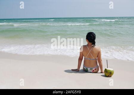 Junge Frau sitzt auf weißem Sand mit Kokosnuss Cocktail neben ihr und die türkisfarbenen Wellen an einem sonnigen Tag Stockfoto