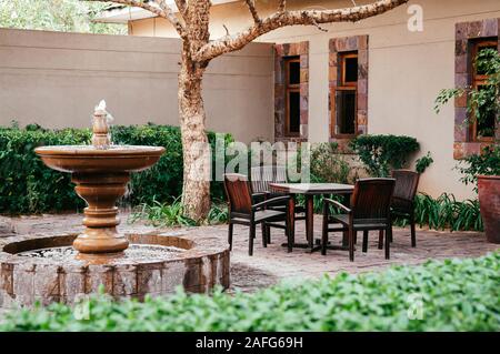 JUN 19, 2011 Serengeti, Tansania - Brunnen und alten hölzernen Stühlen und Tisch in tropischen Garten im Hinterhof unter Baum mit grünen Sträuchern unter Natu Stockfoto