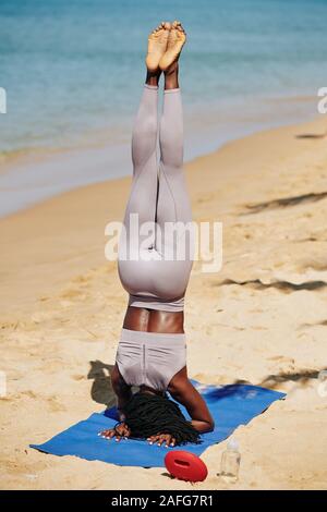 Sportliche junge schwarze Frau Durchführung Kopfstand auf Yoga Matte am Strand, Ansicht von der Rückseite Stockfoto