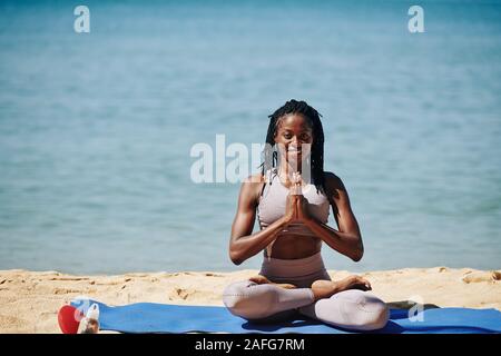 Glücklich lächelnde junge schwarze Frau sitzt auf Yoga Matte auf den Strand und halten die Hände in namaste Geste Stockfoto