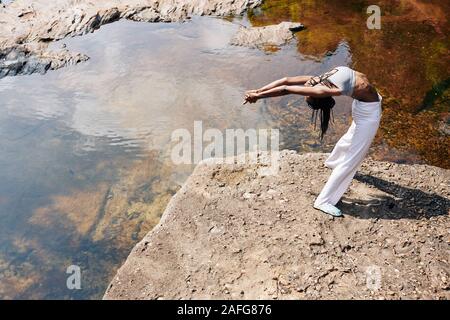 Schwarze junge Frau in Sportswear zurück, biegen beim Stehen auf Rock am Fluss Stockfoto