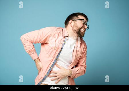 Bärtiger Mann mit Brille Erfahrungen starke Schmerzen im Bauchraum, grinst Zähne und schließt die Augen von Leiden. Blauen Hintergrund. Stockfoto