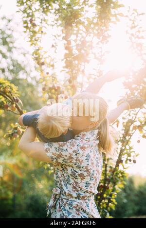 Junge Frau Mutter umarmen und küssen Ihr Baby Sohn im sonnigen Garten. Outdoor Sommer Portrait. Stockfoto