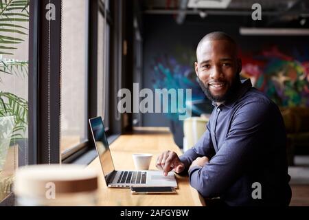 Mitte der erwachsenen männlichen Schwarzen kreative Sitzen durch Fenster in Cafe mit Laptop, drehen und lächelnd in die Kamera Stockfoto