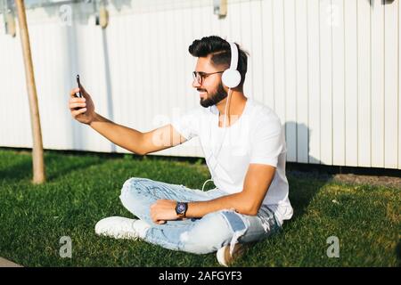 Junge inder Musik vom Handy mit Ohrhörer sitzen auf dem Gras in der Straße. Stockfoto