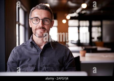 Im mittleren Alter weiße männliche Kreative sitzt in einem Büro lächelnd in die Kamera, Kopf und Schultern, in der Nähe Stockfoto