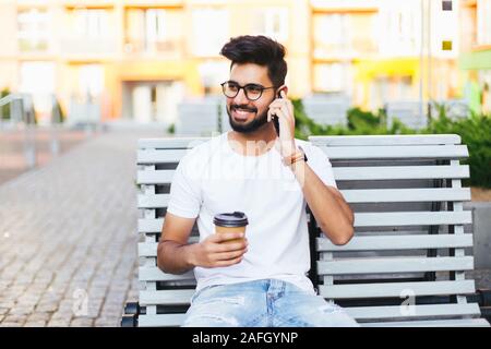Junge indische Mann sitzt auf der Bank, im Gespräch per Telefon, bei Kamera schaut. In stattlicher Mann mit Brille in Weiß Modisches Shirt. Es Arbeitnehmer lächelnd, pos Stockfoto
